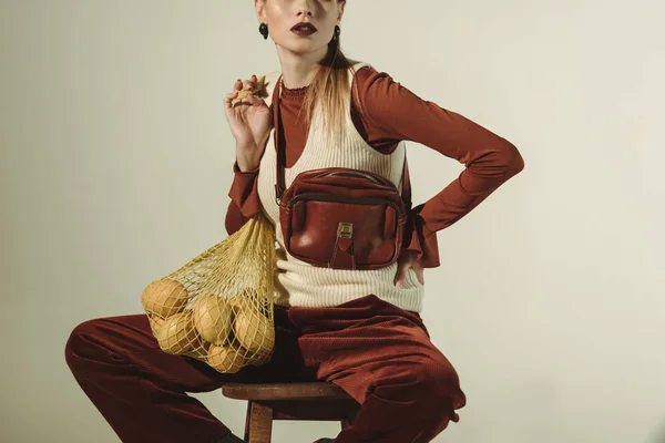 Vue recadrée d'une fille à la mode assise sur un tabouret avec des citrons dans un sac à ficelle isolé sur beige — Photo de stock