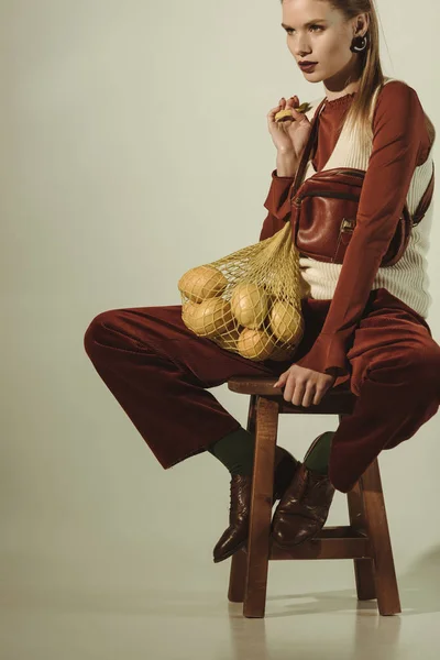 Beautiful stylish woman sitting on stool with lemons in string bag on beige — Stock Photo