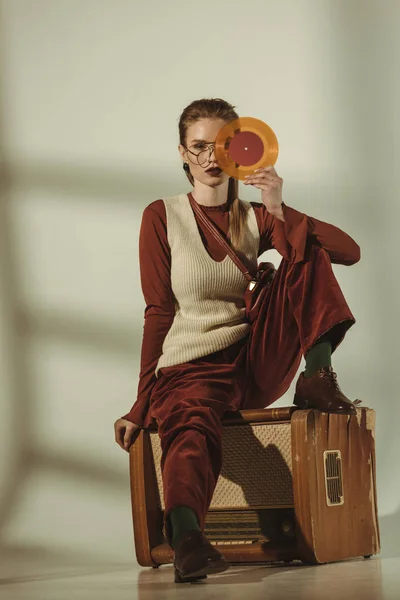 Beautiful young woman holding vinyl record and sitting on vintage tv on beige — Stock Photo