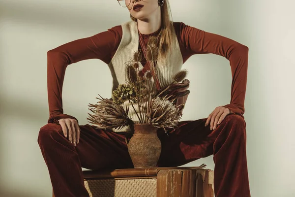 Cropped view of stylish young woman sitting on vintage tv with bouquet of dry flowers on beige — Stock Photo