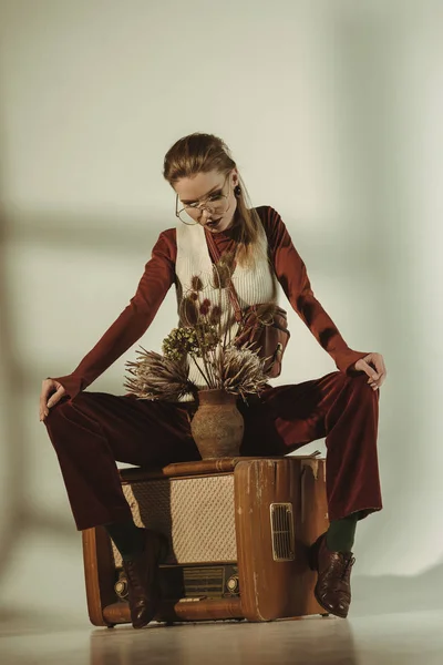 Attractive stylish woman sitting on vintage tv with bouquet of dry flowers on beige — Stock Photo