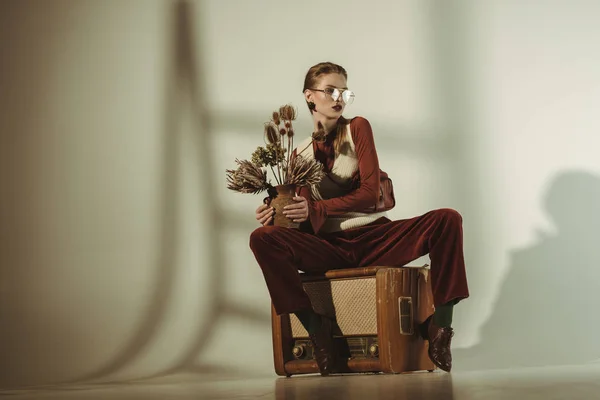 Fashionable girl holding bouquet of dry flowers and sitting on vintage tv on beige — Stock Photo