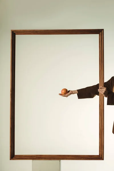 Vista cortada de menina segurando laranja em grande quadro de madeira isolado no bege — Fotografia de Stock