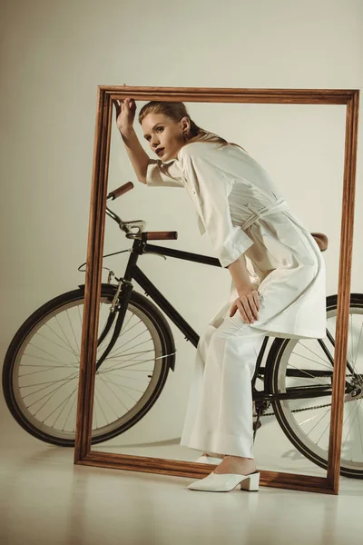 Attractive stylish girl in white outfit posing with bicycle and big wooden frame — Stock Photo