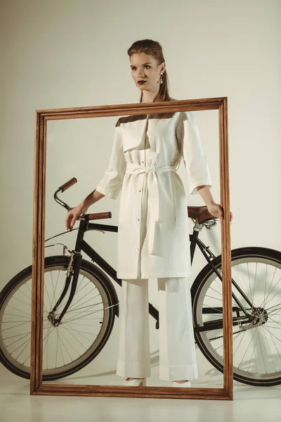 Femme élégante en tenue blanche posant avec grand cadre en bois près du vélo — Photo de stock