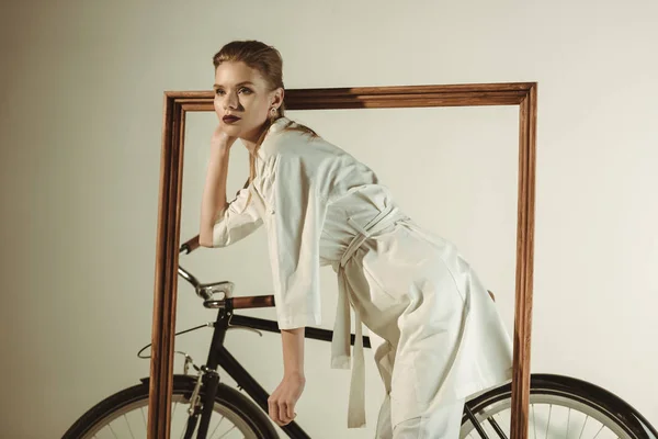 Modèle attrayant à la mode en tenue blanche posant sur un vélo avec un grand cadre en bois — Photo de stock