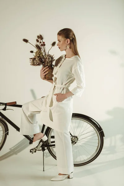 Belle fille élégante posant avec bouquet de fleurs sèches près du vélo — Photo de stock