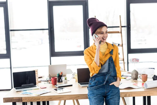 Smiling young female it specialist talking on smartphone near computer desk in loft office — Stock Photo
