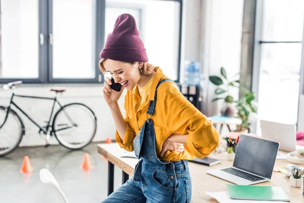 Sorridente giovane femmina è specialista parlando su smartphone vicino a scrivania di computer in ufficio loft — Foto stock