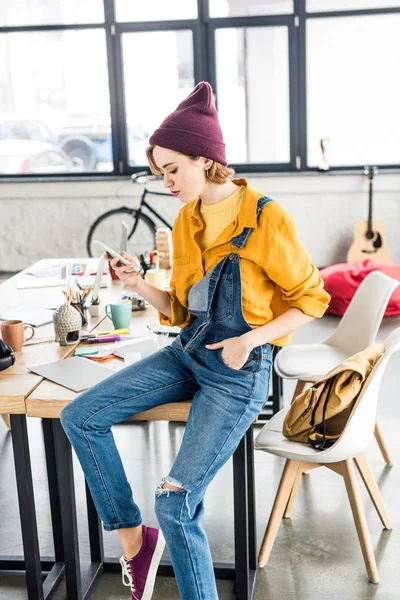 Female designer sitting on table and using smartphone in loft office — Stock Photo