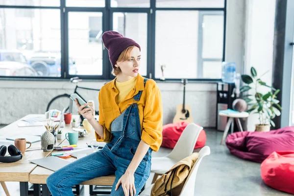 Female designer sitting on table and using smartphone in loft office with copy space — Stock Photo
