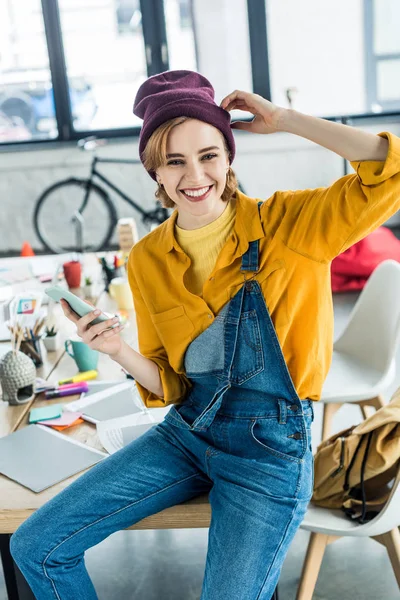 Schöne lächelnde Designerin, die im Loft-Büro auf dem Tisch sitzt und das Smartphone benutzt — Stockfoto