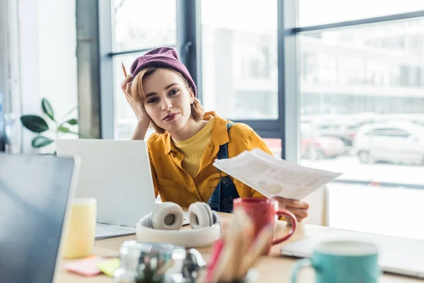 Молодая женщина это специалист, держащий документ и ручку, сидя за компьютерным столом в лофт-офисе — стоковое фото