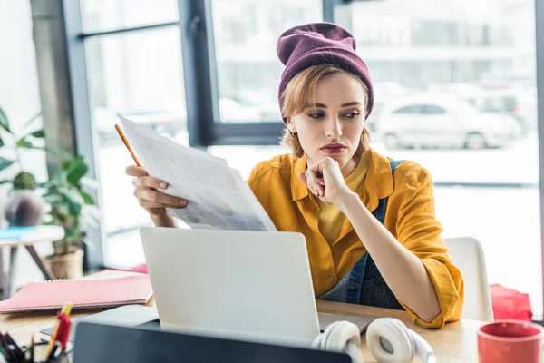 Confused young female it specialist holding document and pen while sitting at computer desk in loft office — Stock Photo