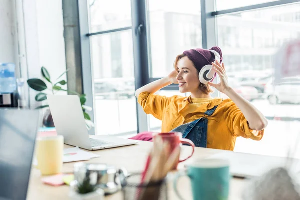 Young female it specialist in headphones listening music while sitting at computer desk in loft office — Stock Photo