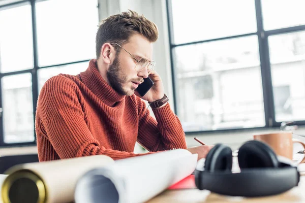 Messa a fuoco selettiva di architetto maschio seduto alla scrivania, parlare su smartphone e lavorare su cianografie in ufficio loft — Foto stock