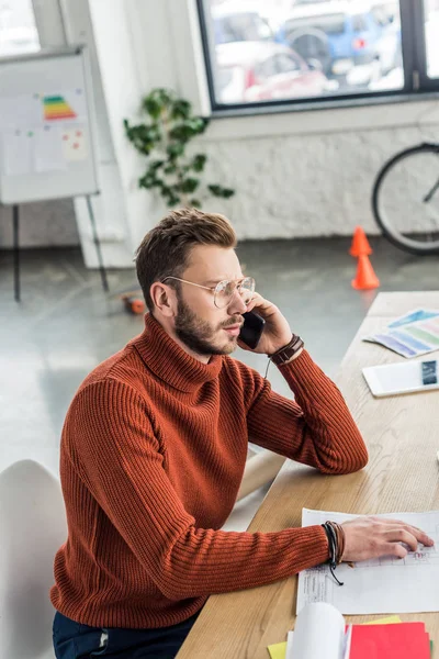 Male architect sitting at desk, talking on smartphone and working on blueprint in loft office — Stock Photo
