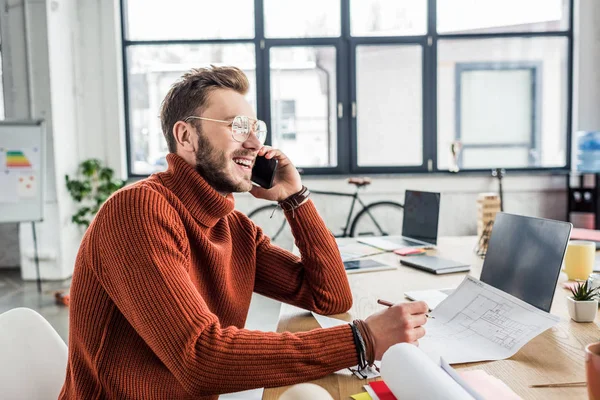 Architetto sorridente seduto alla scrivania, che parla su smartphone e lavora su cianografie in loft office — Foto stock