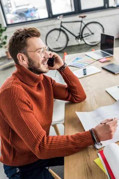 Lächelnder männlicher Architekt sitzt am Schreibtisch, telefoniert mit dem Smartphone und arbeitet im Loftbüro an einem Bauplan — Stockfoto