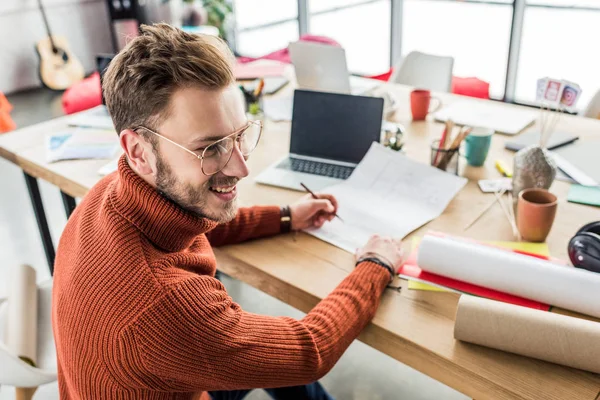 Apuesto arquitecto masculino sonriente sentado en el escritorio y trabajando en planos en la oficina loft - foto de stock