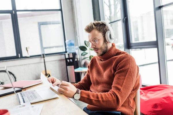 Casual businessman in headphones with document sitting at computer desk in loft office — Stock Photo