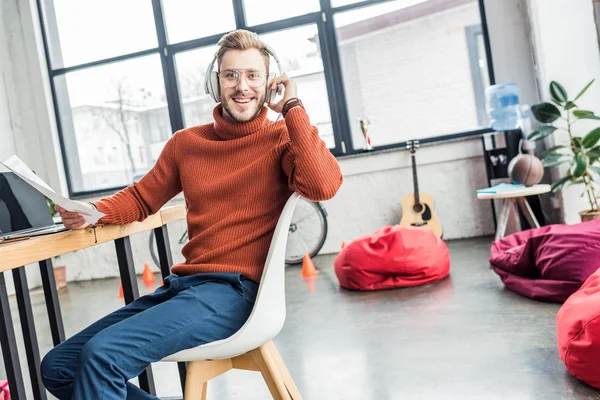 Guapo hombre de negocios casual sentado en el escritorio en los auriculares y trabajando en el documento en la oficina loft - foto de stock