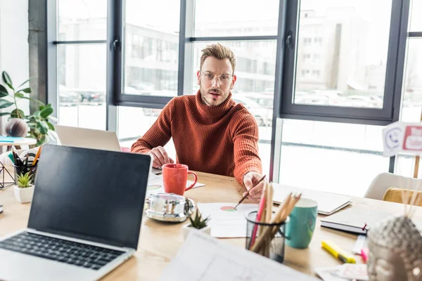 Schöner Gelegenheitsunternehmer sitzt am Computer-Schreibtisch, blickt in die Kamera und arbeitet im Loft-Büro an Dokumenten — Stockfoto