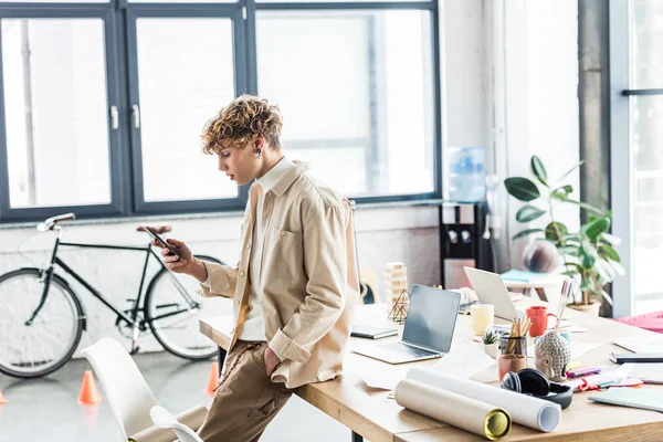 Bello architetto utilizzando smartphone vicino al tavolo con cianografie in loft ufficio — Foto stock