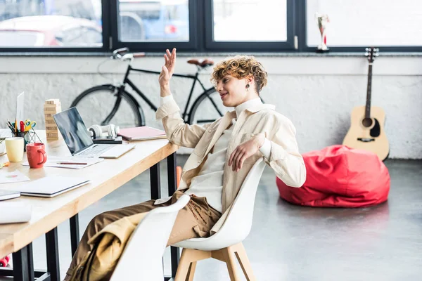 Guapo especialista sentado en el escritorio y haciendo gestos mientras se utiliza el ordenador portátil en la oficina loft - foto de stock