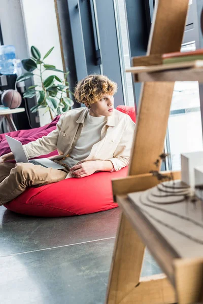 Guapo hombre de negocios casual sentado en la silla de la bolsa de frijol mientras se utiliza el ordenador portátil en la oficina loft - foto de stock