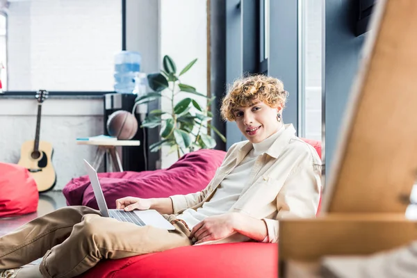 Guapo hombre de negocios casual sentado en la silla de la bolsa de frijol y mirando a la cámara mientras se utiliza el ordenador portátil en la oficina loft - foto de stock