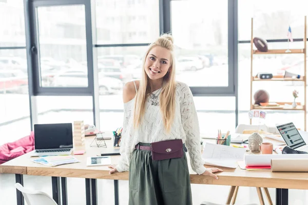 Beautiful female architect looking at camera near table with blueprints and laptop in loft office — Stock Photo