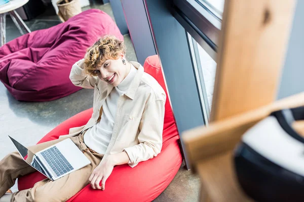 Homem de negócios casual bonito sentado na cadeira de saco de feijão e olhando para a câmera ao usar laptop no escritório loft — Fotografia de Stock