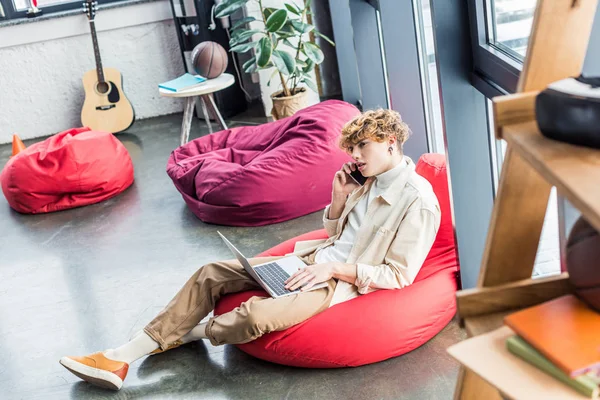 Schöner Gelegenheitsunternehmer sitzt im Sitzsack-Stuhl, während er Laptop benutzt und im Loft-Büro mit dem Smartphone spricht — Stockfoto