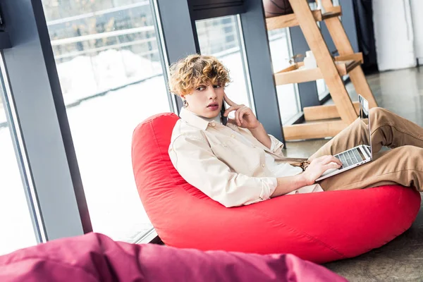 Guapo hombre de negocios casual sentado en la silla de la bolsa de frijol mientras se utiliza el ordenador portátil y hablando en el teléfono inteligente en la oficina loft - foto de stock