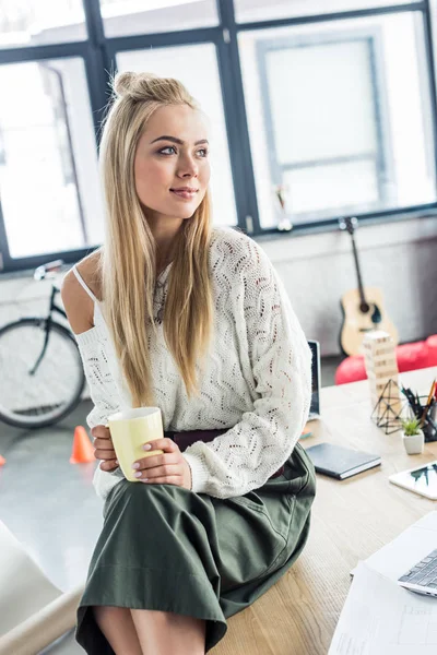 Hermosa mujer de negocios casual sosteniendo la taza de café y mirando hacia otro lado en la oficina loft — Stock Photo