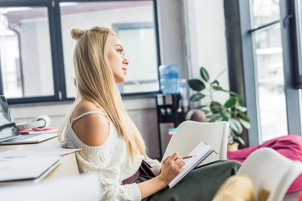 Mujer de negocios casual enfocada sentada y escribiendo en un cuaderno en la oficina del loft - foto de stock