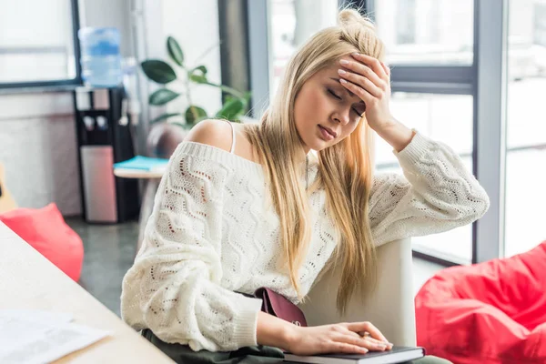 Tired casual businesswoman sitting at desk with notebook and having headache in loft office — Stock Photo