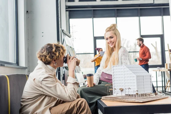 Female and male architects with coffee to go looking at each other while working on house model in loft office — Stock Photo
