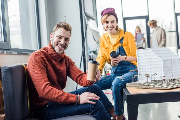 Arquitectos femeninos y masculinos con café para ir mirando a la cámara y trabajando en modelo de casa en la oficina loft - foto de stock