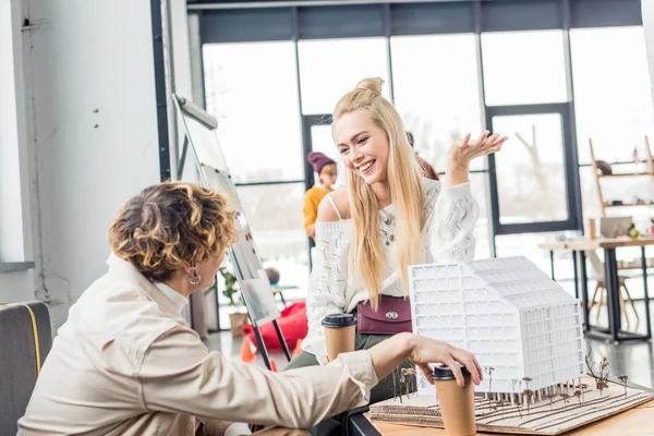 Arquitectos femeninos y masculinos con café para ir mirándose mientras trabajan en el modelo de casa en la oficina loft - foto de stock