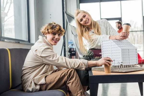 Arquitectos femeninos y masculinos con café para ir mirando a la cámara y trabajando en modelo de casa en la oficina loft - foto de stock