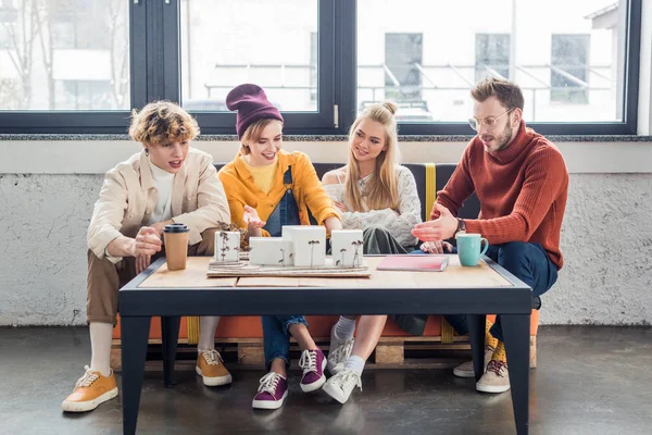 Grupo sonriente de arquitectos femeninos y masculinos sentados en la mesa y trabajando en el modelo de casa en la oficina loft - foto de stock