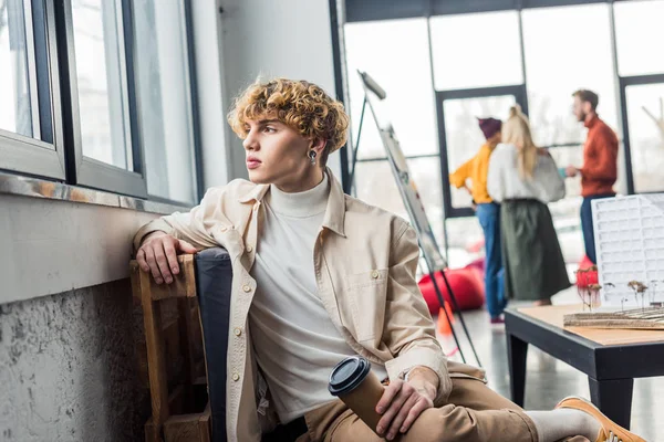 Schöner Mann sitzt mit Kaffee zu gehen im Loft-Büro mit Kollegen im Hintergrund — Stockfoto