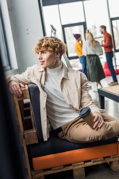 Bel homme assis avec du café pour aller dans le bureau loft avec des collègues sur fond — Photo de stock