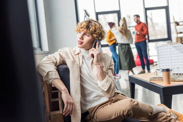 Homem bonito sentado e falando no smartphone no escritório loft com colegas em segundo plano — Fotografia de Stock