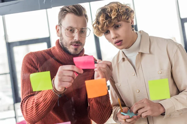 Gutaussehende Gelegenheitsunternehmer kleben bunte Zettel an Glasfenster im Büro — Stockfoto