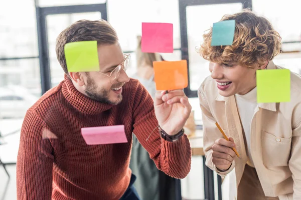 Selective focus of handsome casual businessmen putting colorful sticky notes on glass window in office — Stock Photo