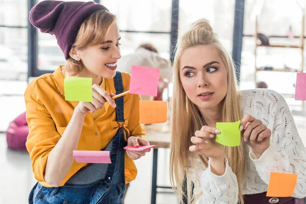 Hermosas mujeres de negocios casuales poniendo notas adhesivas coloridas en la ventana de cristal en la oficina - foto de stock