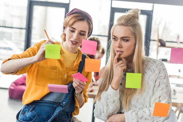 Hermosas mujeres de negocios casuales mirando notas adhesivas de colores en la ventana de cristal en la oficina - foto de stock